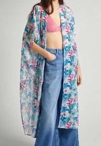 Kvinna som bär en genomskinlig blommig kimono över en rosa bralette och högmidjade vida blå jeans, med handen i fickan, stående mot en enkel bakgrund.