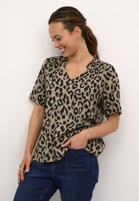 Blusa beige con stampa leopardata, maniche corte, scollatura a V e dettaglio arricciato. Abbinata a jeans in denim blu. Tessuto dalla consistenza liscia.