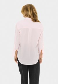Femme avec des cheveux blonds portant un haut à manches longues rose clair et un pantalon sombre, vue de dos sur fond uni.