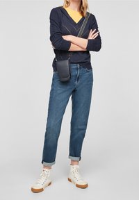 Marinblå v-ringad tröja över en gul skjorta, kombinerat med uppvikta blå jeans och vita högstövliga sneakers med bruna sulor. Svart axelremsväska.