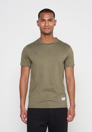 Mann steht frontal, trägt ein olivgrünes kurzärmeliges T-Shirt und schwarze Hosen vor einem einfarbigen hellen Hintergrund.