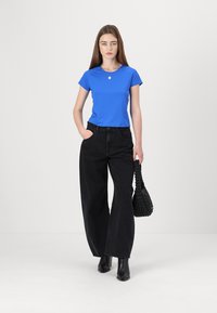 Top blu corto a maniche corte abbinato a jeans neri a vita alta e a gamba larga. Il modello tiene in mano una borsa nera con trama e indossa stivali alla caviglia.