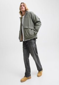 Wodoodporna kurtka w kolorze oliwkowej zieleni z zamkiem z przodu i logo na piersi, w połączeniu z szarymi dżinsami i beżowymi butami. Gładka tekstura, luźny krój i minimalistyczny design.