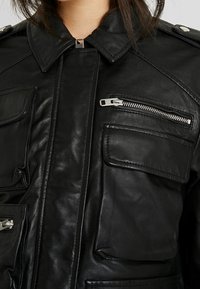 Černá kožená bunda s límcem, několika kapsami a zipovými detaily. Hladká textura s viditelnými švy.