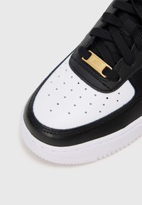 Sapatilha em preto e branco com parte superior em pele, bico perfurado e detalhe em hardware dourado. Design clássico com sola de borracha e fecho de atacadores.