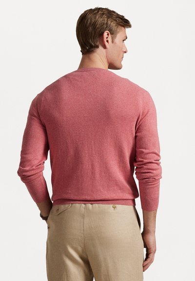 Vyrų raudonas megztinis ilgomis rankovėmis ir apvalia iškirpte, pagamintas iš minkštos medžiagos. Dėvimas ant šviesių smėlio spalvos kelnių, demonstruojančių lygų audinį.