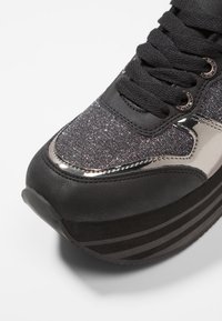 Musta urheilukengät metallisilla yksityiskohdilla, kimaltavasta tekstiilistä valmistettu päällinen, pyöreä kärki, paksu musta pohja, jossa on vaakasuoria raitoja, ja teksturoitu nauha.