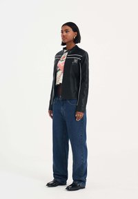 Svart fuskläderjacka med vit sömnad, kombinerad med lösa blå jeans. Har en dragkedja fram och en grafisk t-shirt under.