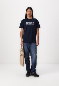 Marineblauwe katoenen t-shirt met het "TOMMY" logo in wit en rood. Gekenmerkt door blauwe jeans en het dragen van een beige tas. Zwarte schoenen maken de outfit compleet.