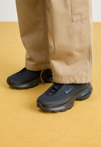 Chaussures de sport noires avec un upper en tissu texturé, bout arrondi et semelles en caoutchouc proéminentes. Assorties à un pantalon cargo ample de couleur beige.