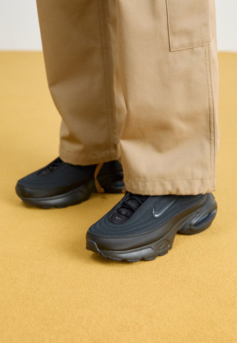 Chaussures de sport noires avec un upper en tissu texturé, bout arrondi et semelles en caoutchouc proéminentes. Assorties à un pantalon cargo ample de couleur beige.