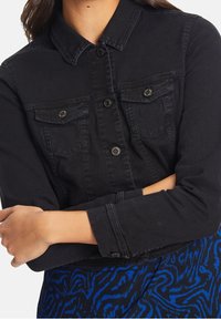 Svart denimjacka med långa ärmar, spetsig krage, bröstfickor och metallknappar. Har en figursydd design och en något slitna yta.