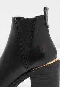 Botín negro que presenta un upper de cuero, costados elásticos y un tacón de madera texturizado con un acento dorado en la base.