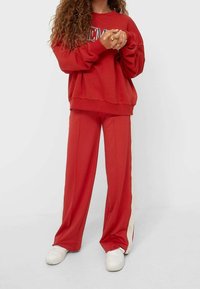 Rode trui met grote letters, gecombineerd met rode wijde broek met zijstrepen. Afgemaakt met witte sneakers. Casual outfit.