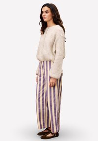 Vrouw die zijwaarts staat, gekleed in een roomkleurige gebreide trui, beige broek met brede pijpen en verticale paarse strepen, en donkerbruine Mary Jane schoenen.
