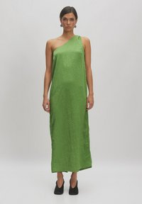 Ein einseitiges grünes Kleid aus einem zerknitterten Stoff mit einer langen, geraden Silhouette. Getragen mit schwarzen Absatzschuhen und Ohrringen.