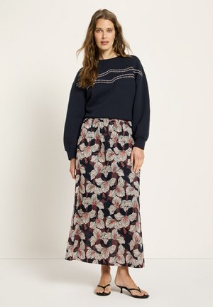 Frau trägt einen marineblauen Pullover mit gesticktem Muster und einen langen schwarzen Rock mit großem weißem und rotem Blumenmuster, steht in Sandalen.