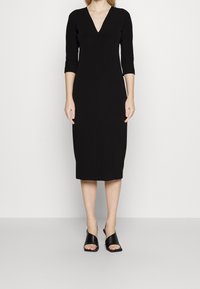 Svart midi-klänning med trekvartslånga ärmar och V-ringning. Slät textur, figursydd silhuett och knälång design. Bärs med svarta öppna tå-skor.
