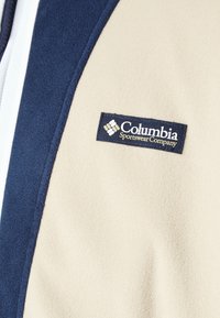 Gros plan sur une veste en polaire beige et marine avec un patch logo de Columbia Sportswear Company sur la poitrine.