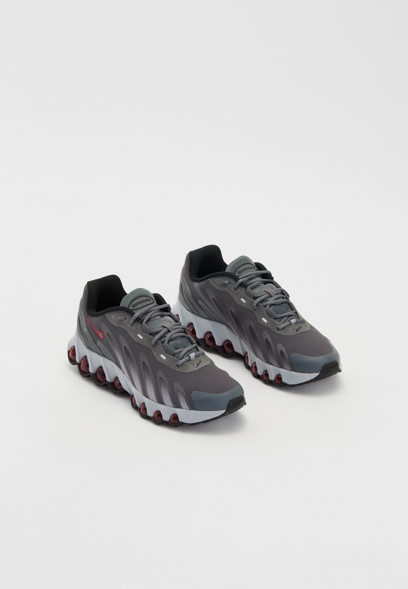 Zapatillas deportivas grises con parte superior de malla y sintético, que presentan acentos rojos, una suela redondeada y un acolchado grueso para mayor soporte.
