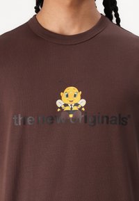 Brun bomull t-skjorte med grafikk av en tegneseriebi og teksten "de nye originalene" i svart trykk under designet.
