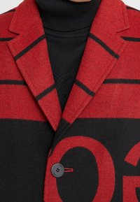 Czerwono-czarny płaszcz z mieszanki wełny, z wyciętym kołnierzem, dużymi klapami i zapięciem na guziki z ozdobnym przeszyciem. Widoczny wzór w paski.