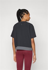 Top negro de manga corta con un diseño ligeramente recortado, escote redondeado y dobladillo gris. Dos botones en la parte posterior del escote para el cierre.