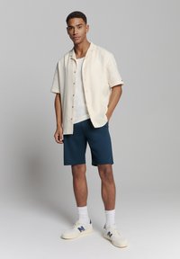 Lichtbeige, kortemouw overhemd met knoopjes, witte tanktop, donkerblauwe shorts en witte sneakers met marineblauwe accenten, tegen een neutrale achtergrond.