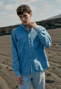 Svetlo modra postavna srajca iz_DENIM_ materiala, z ovratnikom in dolgimi rokavi, v kombinaciji s svetlo modrimi kavbojkami na teksturirani pokrajini.