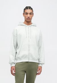 Šviesiai pilka zip-up gobtuvė su kišenėmis kangaroo stiliumi ir apyrankėmis, vilkima ant žalių treningų. Lygus paviršius, kasdieniškas dizainas.