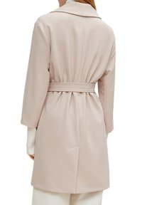Ein hellrosa Trenchcoat mit tailliertem Gürtel, breiten Revers und langen Ärmeln. Der Stoff wirkt glatt und leicht strukturiert.