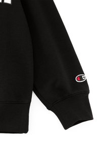 Sudadera negra con una textura de punto estructurado, que presenta un puño acanalado y un pequeño logo bordado en rojo y blanco en la manga.