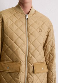 Veste bomber matelassée beige avec col côtelé, fermeture éclair argentée et deux poches plaquées à l'avant. Présente un logo discret sur la poitrine.