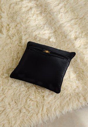 Black velvet cushion with a zip closure and gold hardware accent, placed on a fluffy cream rug. Square shape with a smooth texture.