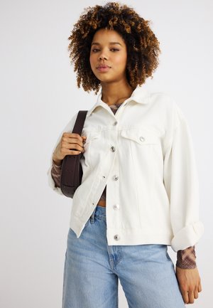 Młoda kobieta o kręconych włosach, ubrana w białą zapinaną kurtkę, niebieskie dżinsy i niosąca ciemnobrązową torebkę na ramię.