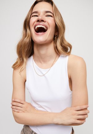 Top blanco sin mangas hecho de tela suave con escote redondo. Usado con un collar delgado de cadena dorada, con cabello ondulado y brazos cruzados.