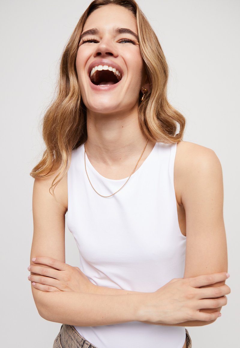 Top blanco sin mangas hecho de tela suave con escote redondo. Usado con un collar delgado de cadena dorada, con cabello ondulado y brazos cruzados.