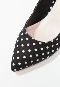 Black pointed-toe shoe with white polka dots and a light beige inner lining, shown from a top close-up angle.