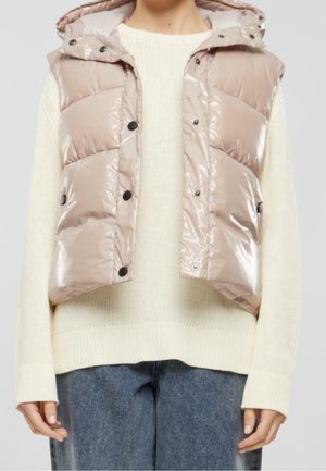 Personne portant une veste matelassée beige brillante par-dessus un pull en tricot crème et un jean bleu, debout devant un fond uni.