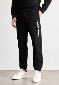 Juodi džinsiniai kelnių tipo šortai, pagaminti iš medvilnės mišinio, su balta Calvin Klein logo vertikaliai kairėje kojoje, su elastingais rankogaliais.