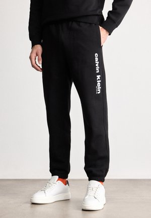 Czarne dresy wykonane z mieszanki bawełny, z białym logo Calvin Klein umieszczonym pionowo wzdłuż lewej nogawki, z elastycznymi mankietami.