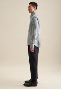 Camicia a quadri bianca e blu; maniche lunghe con polsini, abbinata a pantaloni blu scuro e scarpe nere. Il materiale sembra tessuto.