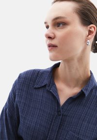 Marinblå, strukturerad skjorta med krage, knappstängning och bröstficka. Modellen bär silveröra med abstrakt form.