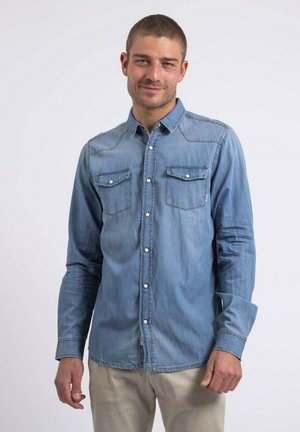 Chemise en denim bleu clair avec un col boutonné, deux poches poitrine et des boutons blancs. Elle présente des manches longues et un ourlet droit.