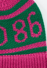 Close-up of knitted fabric with pink and green yarn, featuring the year "1986" in pink on a green background.