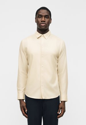 Άνδρας που φοράει μπεζ μακρυμάνικο πουκάμισο με κουμπιά και μαύρο παντελόνι, στέκεται όρθιος μπροστά σε ένα απλό φωτεινό φόντο.