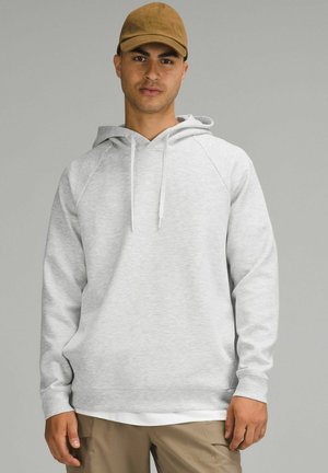 Haut en molleton gris clair avec cordon de serrage et poche kangourou. Il présente une texture douce et des manches raglan, associé à une casquette beige.