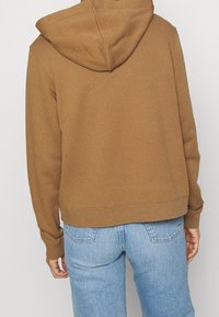 Felpa marrone con cappuccio, tessuto morbido, polsini e orlo a coste, abbinata a jeans azzurri. Presenta una vestibilità comoda e un design semplice.