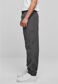 Seitenansicht einer Person, die dunkelgraue Jogginghosen mit elastischen Knöcheln und weiße Sneakers mit Schnürsenkeln vor einem grauen Hintergrund trägt.