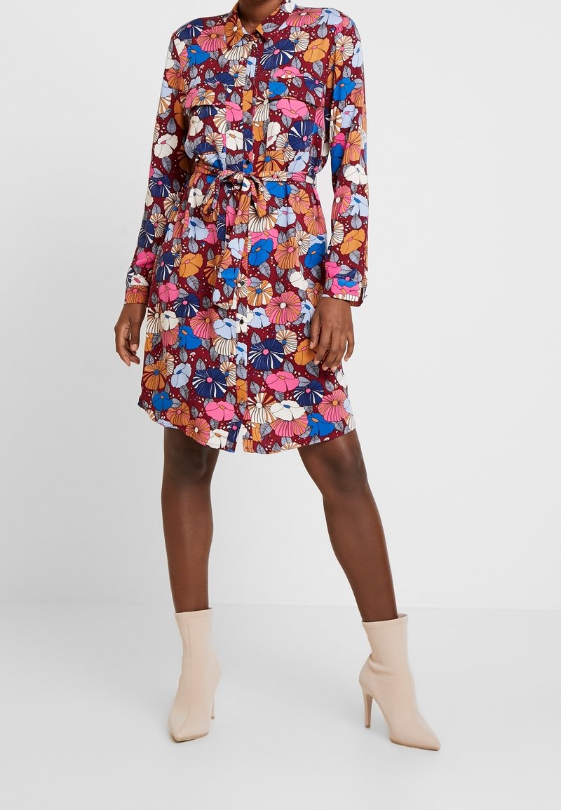 Femme portant une robe chemise boutonnée à manches longues avec un motif floral coloré et une ceinture à la taille, associée à des bottines beiges à talons hauts.